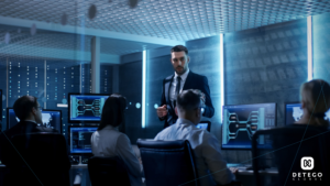 A young white man in a suit speaks to a team of 4 other people sitting in front of computer screens