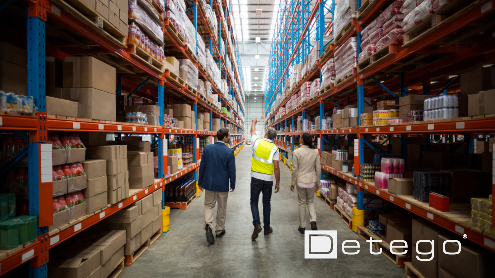 A worker in a yellow vest leads a businessman and a businesswoman in suits between tall shelves in a warehouse