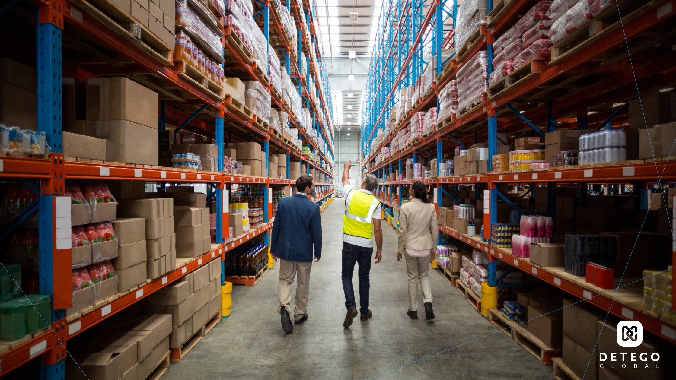 Warehouse workers walk down an aisle between two tall shelves stacked with cardboard boxes