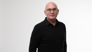 A bald Caucasian man wearing glasses and a black shirt standing against a white background