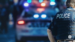 A uniformed male police officer stands in the foreground in front of a police car with flashing red and blue lights