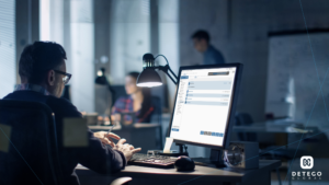 A man sits with his back to the camera, working at a brightly lit computer screen in an office