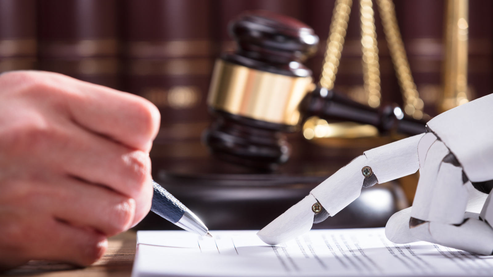 A human hand holding a pen and a robot hand scan a paper document in front of a judge's gavel and the scales of justice