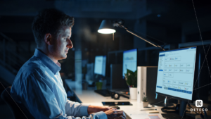 A white man sits in front of a computer at a brightly lit desk in a dark room