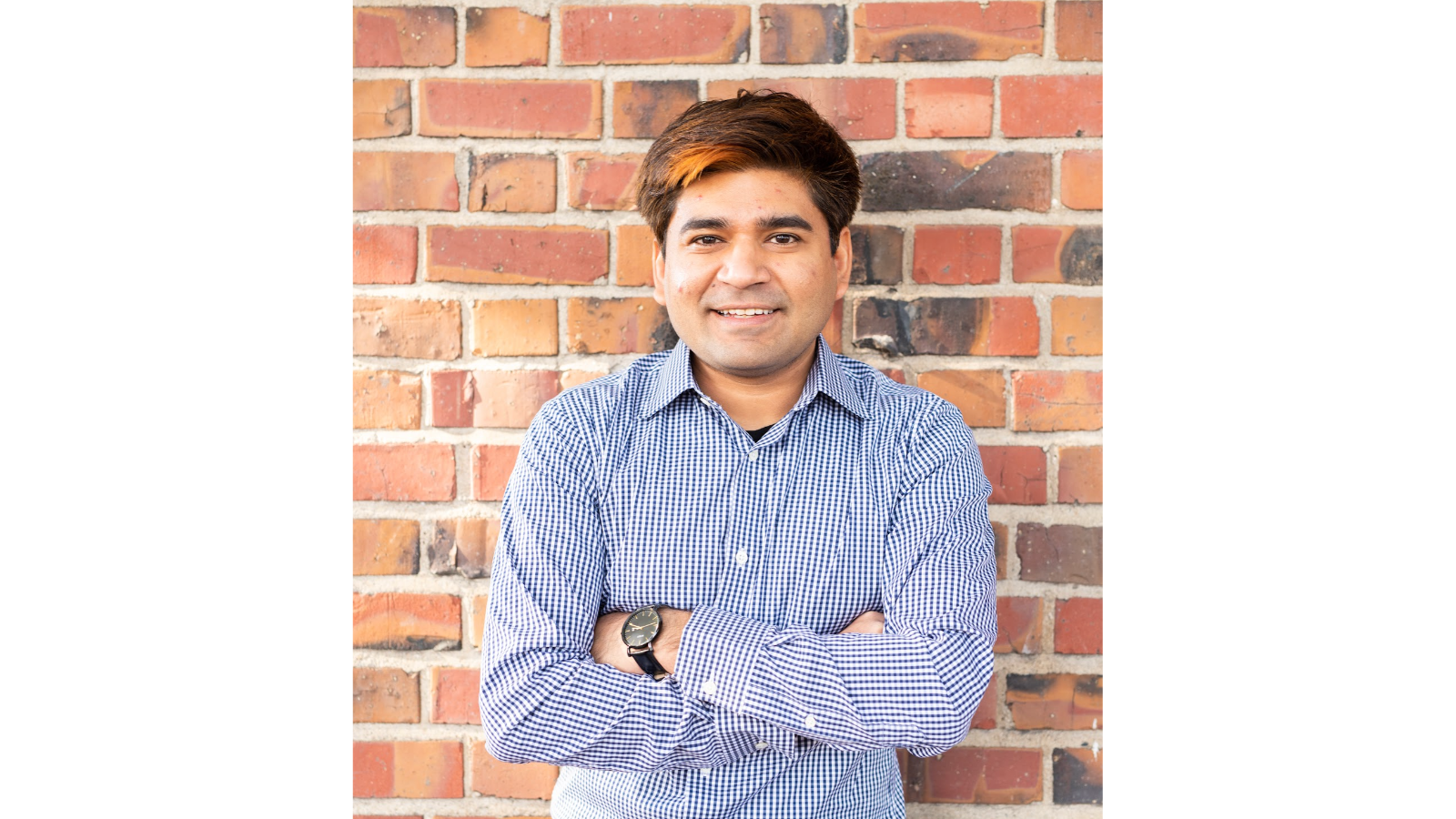 A man in a blue shirt stands with arms crossed against a brick wall backdrop